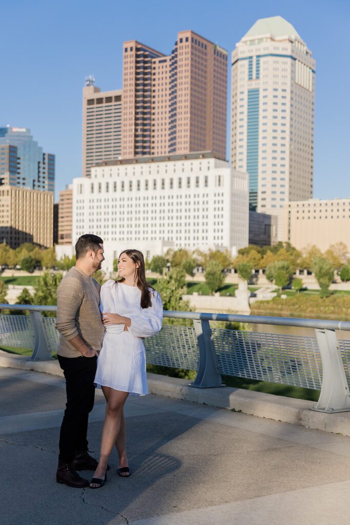 Downtown Columbus Engagement Photo on Scioto Mile – A couple embraces with the Columbus skyline and Scioto River in the background at the Scioto Mile.